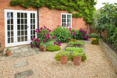 Charming garden patio with gravel path, potted plants, and vibrant flowers against a red brick exterior with white French doors.